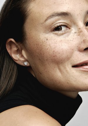 Silberne, herzförmige Steckerohrringe an einer Frau mit Sommersprossen, die ein schwarzes hochgeschlossenes Oberteil trägt. Glatte Textur, einfaches Design, kleine Größe.