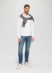 Witte lange mouwen knoopsluiting shirt, lichtblauwe denim jeans, grijze gebreide trui over de schouders, en witte sneakers met grijze accenten.