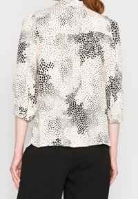 Blusa chiara a fantasia con pois neri, cierre a bottoni sul retro e maniche a 3/4. Il tessuto sembra morbido con un leggero lucido.