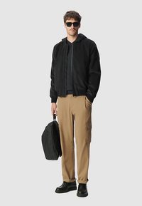 Giacca bomber nera con zip, strato interno grigio, pantaloni cargo color cognac e stivali neri. Tiene uno zaino nero. In piedi contro uno sfondo grigio chiaro.
