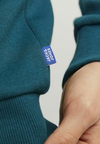 Felpa verde acqua con polsini a costine, dotata di un piccolo etichetta blu con il testo "JACK & JONES". Il tessuto è liscio, con una leggera trama.