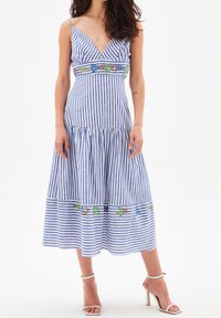 Wanita mengenakan gaun midi bergaris vertikal biru dan putih dengan sulaman bunga di pinggang dan hem, dipadukan dengan sandal hak tinggi putih.