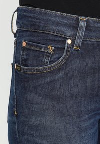 Jeans de mezclilla en azul oscuro con una textura sutil. Presentan bolsillos tradicionales, remaches de cobre y costuras amarillas a lo largo de las costuras.