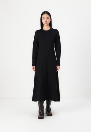 Vestido de algodón negro de manga larga con cuello redondo y dobladillo acampanado, combinado con botas de tobillo marrón oscuro. Textura suave, diseño minimalista.