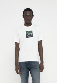 Άσπρο βαμβακερό μπλουζάκι με τετράγωνο πράσινο και ναυτικό λογότυπο στην πρόσοψη, σε συνδυασμό με μπλε τζιν τζιν. Απλός στρόγγυλος λαιμός.