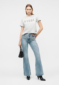 Femme portant un t-shirt Ksubi gris clair, un jean taille haute bleu orné de décorations, des bottes à talons noires, tenant un sac à main noir clouté.
