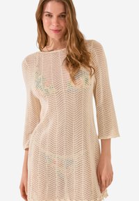 Abito maglione in crema con un motivo a tessitura aperta, maniche a 3/4 e orlo a scallop. Presenta sottili accenti colorati.