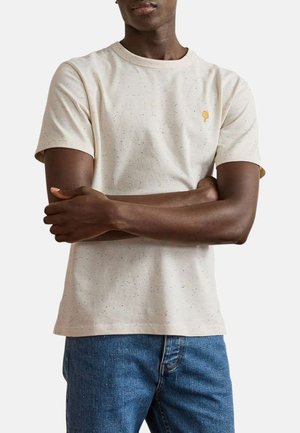Homme les bras croisés portant un t-shirt à manches courtes beige éclaboussé et un jean bleu, sur un fond uni.