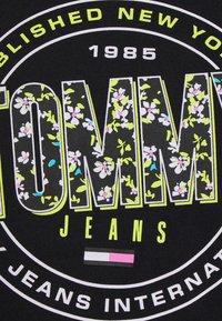 Czarna tkanina z dużym, wyraźnym napisem "TOMMY JEANS" z wzorami kwiatowymi, akcentowana neonowym żółtym, różowym i niebieskim. Obecny okrągły wzór logo.
