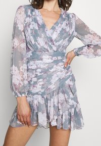 Blommig klänning i genomskinlig ljusblå chiffon, med långa ärmar, omslagsdesign, plisserade detaljer och en volangkant dekorerad med lila och vita blommor.