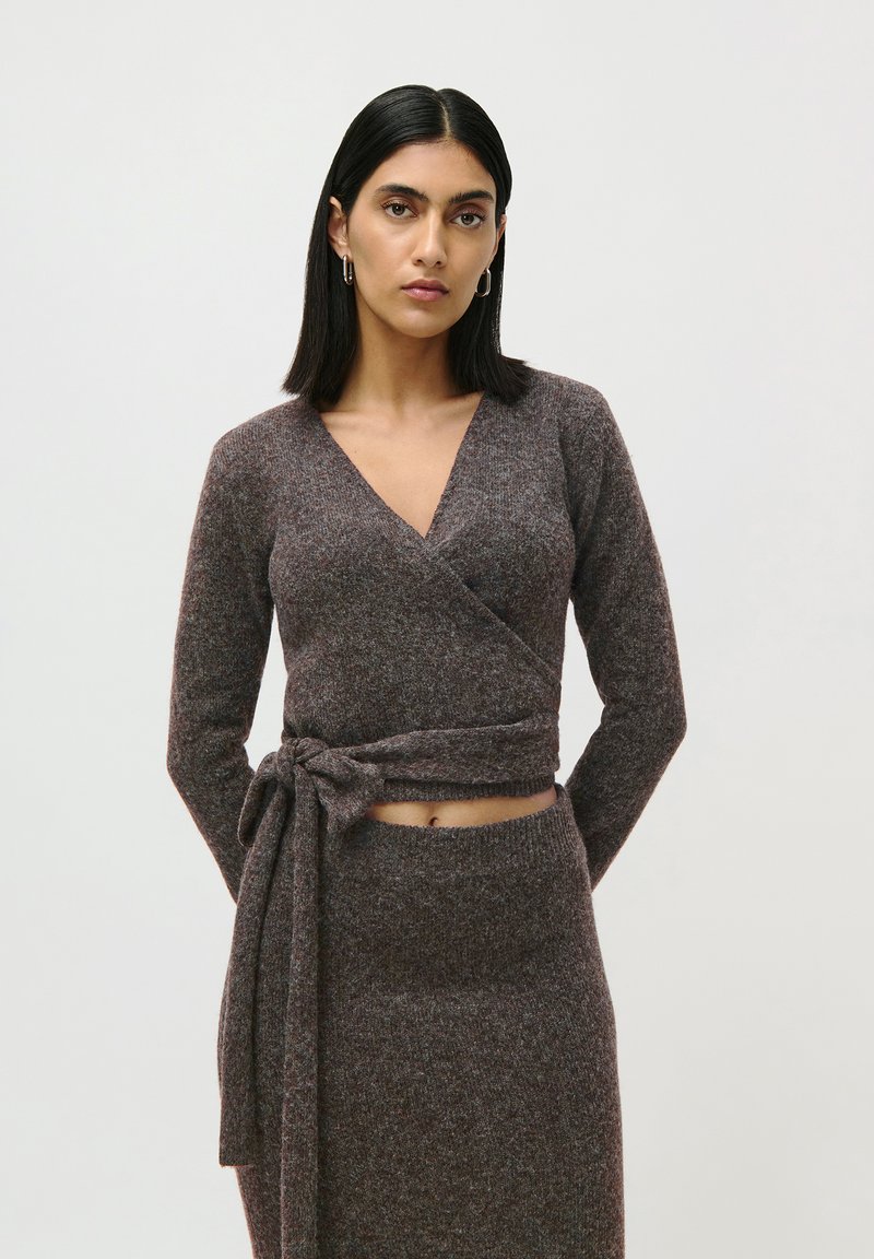 Femme jeune avec des cheveux noirs raides portant un pull enveloppant marron noué à la taille et une jupe en tricot assortie sur fond uni.