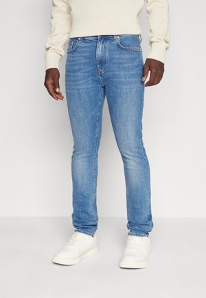 Mann trägt hellblaue Slim-Fit-Jeans, weiße Sneaker und einen cremefarbenen Rippstrickpullover und steht vor weißem Hintergrund.