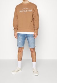 Sudadera marrón con texto blanco, combinada con pantalones cortos de mezclilla azul claro con dobladillos deshilachados y zapatillas beige con detalles decorativos.
