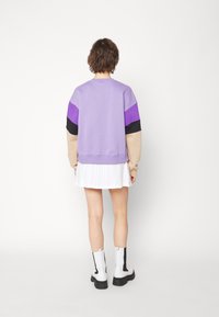 Sudadera de forro polar morada con mangas en bloques de color en morado oscuro y negro, combinada con una falda blanca plisada y botas blancas de tobillo.