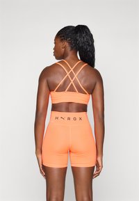 Oransje trenings-BH med kryssede stropper og matchende høyt midjebukser, med svart "HYROX" bokstaver på midjebåndet. Glatt tekstur.
