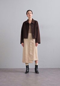 Barna gyapjú cropped dzseki gallérral és gombokkal, bézs térdígyérő ruhával párosítva. Fekete bokacsizmák egészítik ki az öltözéket.