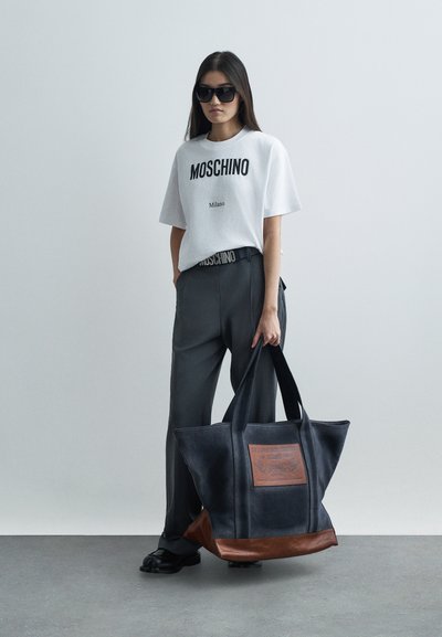 Γυναίκα που φοράει oversize πουκάμισο και παντελόνι Moschino, κρατώντας μεγάλη μαύρη και καφέ τσάντα, στέκεται μπροστά σε απλό φωτεινό φόντο.