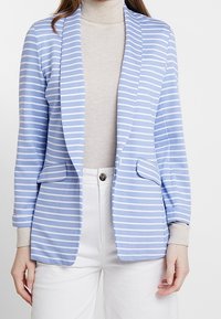 Blazer a righe in bleu chiaro e bianco, con collo sciallato, due tasche frontali e una vestibilità relax. Il tessuto sembra morbido e testurizzato.