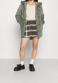 Kvinna iklädd en grå pälsliknande jacka med huva, beige topp, rutig kjol, vita strumpor och svarta tofflor med fransar, står med händerna i fickorna.