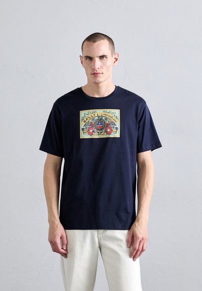 T-shirt en coton bleu marine avec un grand imprimé graphique comportant du texte et des motifs. Col rond simple et manches courtes, associé à un pantalon de couleur claire.