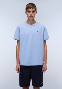 Light blue cotton t-shirt with short sleeves, round neck, and small embroidered logo on the chest. Worn with dark navy shorts.