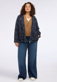 Marinblå huva jacka med vit rutigt mönster, dragkedjestängning och två framfickor över en brun cardigan och blå vida jeans.