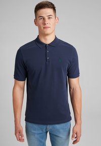 Polo de color azul marino con una tapeta de dos botones, mangas cortas y un logo bordado en verde en el lado izquierdo del pecho. Textura de tela suave.