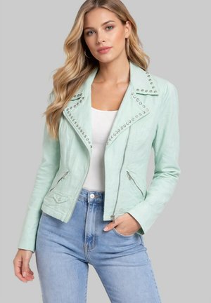 Jeune femme aux longs cheveux blonds portant une veste en cuir verte menthe cloutée, un haut blanc et un jean bleu clair sur fond gris.