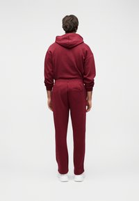 Egy férfi, aki bordó kapucnis pulóvert és melegítőnadrágot visel, háttal áll, fehér sportcipőben, egyszínű fehér háttér előtt.