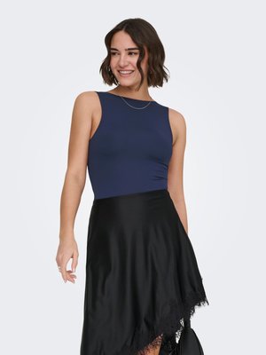 Vrouw met kort donker haar lacht, draagt een mouwloze marineblauwe top en een asymmetrische zwarte rok met kanten rand, tegen een effen achtergrond.