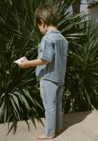 Outfit in denim composto da una maglietta a maniche corte e pantaloni lunghi di colore blu chiaro. Il bambino è scalzo e tiene un oggetto di carta, in piedi di fronte a un'area verde.