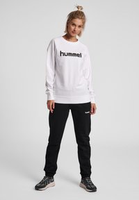 Felpa bianca a maniche lunghe con logo "hummel" nero, abbinata a pantaloni jogger neri con dettagli del logo, e scarpe da ginnastica grigio e nere.