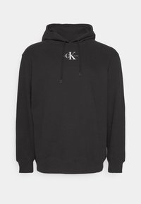 Čierna mikina s kapucňou vyrobená z mäkkej látky, s bielym logom na prednej strane. Má kapucňu so sťahovacou šnúrkou a rebrované manžety a spodný lem.