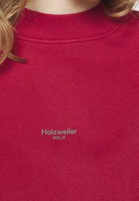 Pull rouge avec col côtelé et logo gris "Holzweiler OSLO" centré sur la poitrine, porté par une personne aux cheveux blonds.