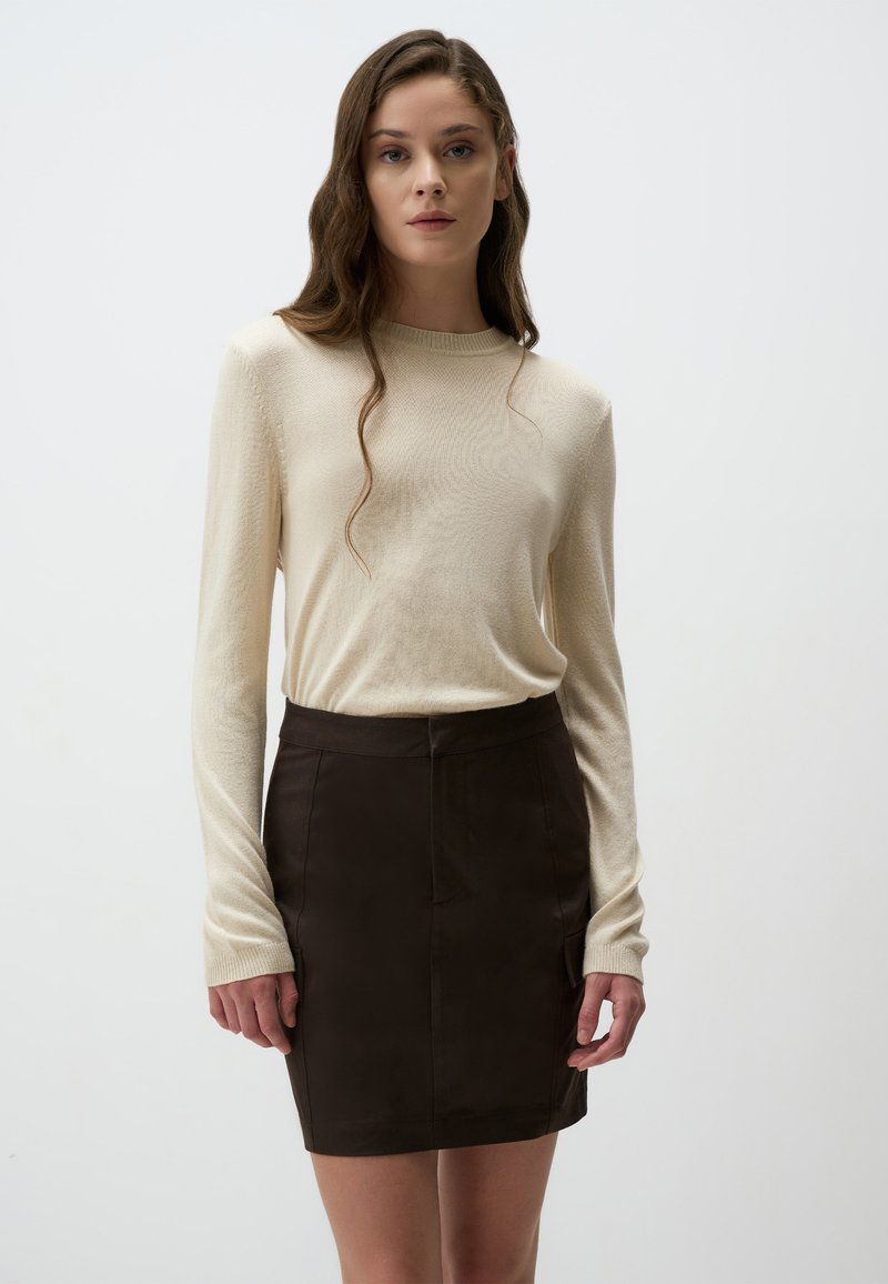 Beige langwerpige trui met lange mouwen, gecombineerd met een aansluitende bruine minirok. De trui heeft een geribbelde textuur en de rok heeft zakken aan de voorkant.