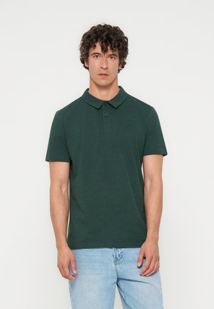 Jeune homme aux cheveux bouclés portant un polo vert foncé à manches courtes et un jean bleu clair, debout sur un fond blanc uni.