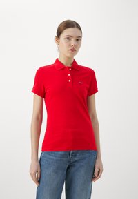 Camiseta polo de algodón roja con hebilla de dos botones y mangas cortas, que presenta un pequeño logo en el lado izquierdo del pecho. Combinada con jeans de mezclilla azul.