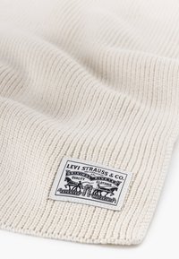 Tissu tricoté côtelé couleur crème avec une finition texturée, présentant une étiquette rectangulaire noir et blanc avec le nom de la marque et des graphiques.