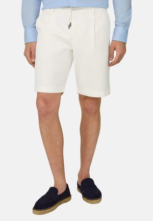Mann trägt weiße maßgeschneiderte Shorts, hellblaues langärmeliges Hemd und marineblaue Slip-On-Schuhe mit gewebten Sohlen, steht vor schlichter Hintergrund.