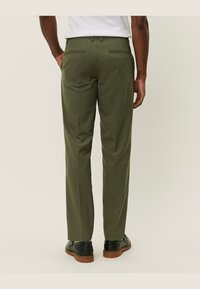 Pantalon vert olive avec coupe ajustée, comportant deux poches arrière, et associé à des chaussures noires sur un fond clair.