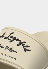 Μπεζ λουράκι σανδάλας με ανάγλυφο μαύρο καλλιγραφικό κείμενο «Karl Lagerfeld Côte D'Azur RIVIERA» σε λευκό φόντο.