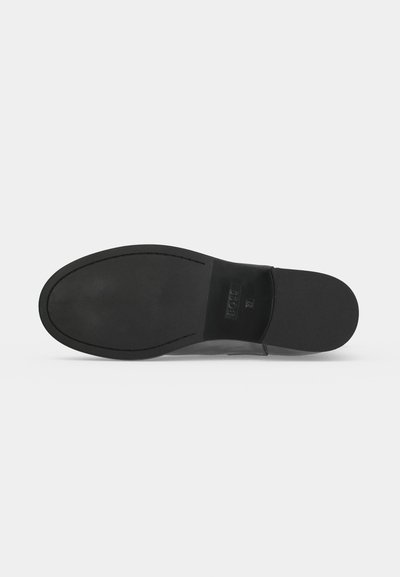 Semelle d'une chaussure de ville en cuir noir, vue de dessous, montrant une texture lisse et des estampilles de marque discrètes près du talon sur un fond uni.