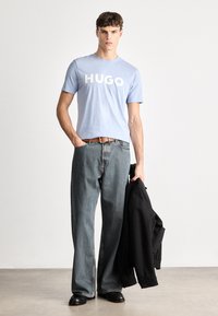 Ανοιχτού μπλε βαμβακερό μπλουζάκι με λογότυπο "HUGO", συνδυασμένο με φαρδύ τζιν παντελόνι σε σκούρο χρώμα. Μαύρο μπουφάν ριγμένο στον βραχίονα. Μαύρα παπούτσια.