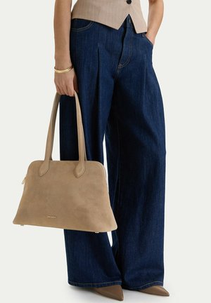 Person trägt dunkelblaue Jeans mit weitem Bein und taupefarbene spitze Schuhe, hält eine passende taupefarbene Handtasche mit doppeltem Griff.