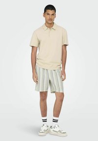 Beige polo krekls ar teksturētu audumu, apvienots ar gaišiem svītraina šortiem zaļā un baltā krāsā, un baltiem sporta apaviem ar pelēkiem akcentiem.