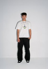 Biała koszulka z kolorowym nadrukiem na środku, w połączeniu z czarnymi spodniami typu cargo i czarnymi sneakersami. Prosty, casualowy wygląd.