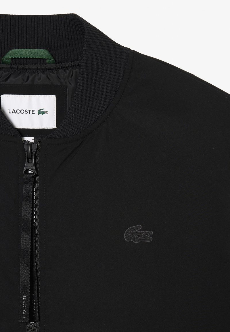 Veste bomber noire avec col côtelé, fermeture éclair et logo discret Lacoste. Texture lisse et design épuré.