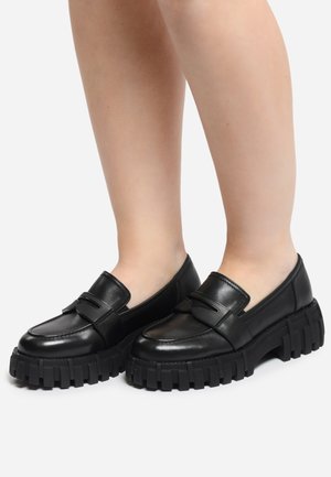 Mocassins en cuir noir avec des semelles en caoutchouc noir chunky, présentant un accent rectangulaire et une finition lisse. Le design est épuré avec un minimum de coutures.