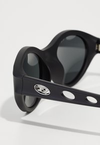 Lunettes de soleil noires au fini mat, de forme ronde, avec des verres sombres et un logo argenté sur la branche comportant des découpes circulaires.