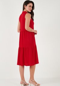Red sleeveless dress with a tiered design, soft fabric, knee-length, and subtle texture. Paired with light-colored strappy sandals.